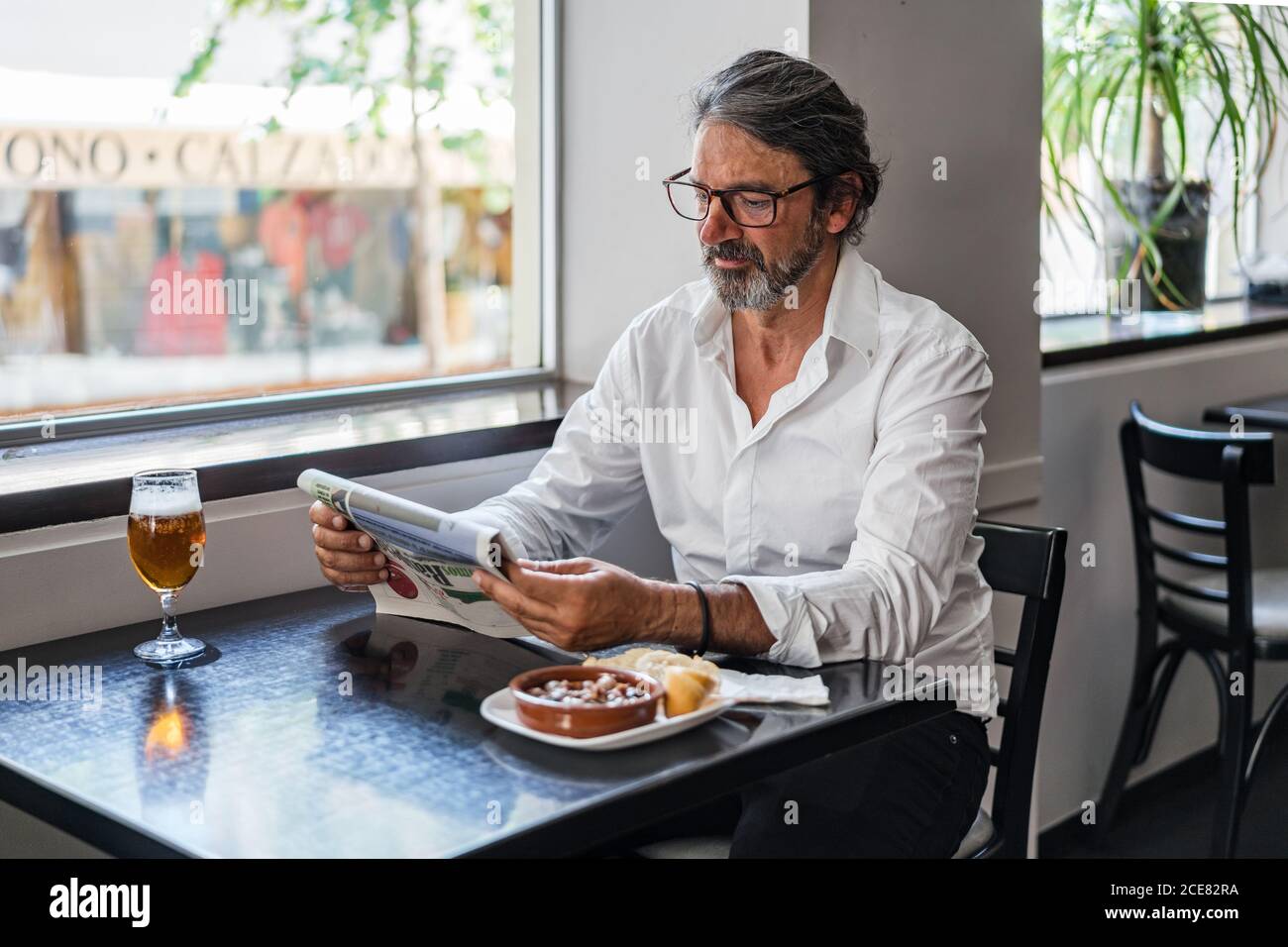 Positive ethnic bearded aged man sitting at table with of glass of foamy beer and reading newspaper near window Stock Photo