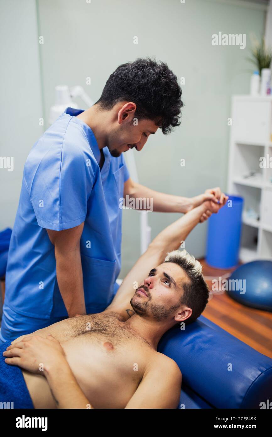 Osteopath in blue uniform examining arm of slim male patient with dyed hair lying on examination table in clinic Stock Photo