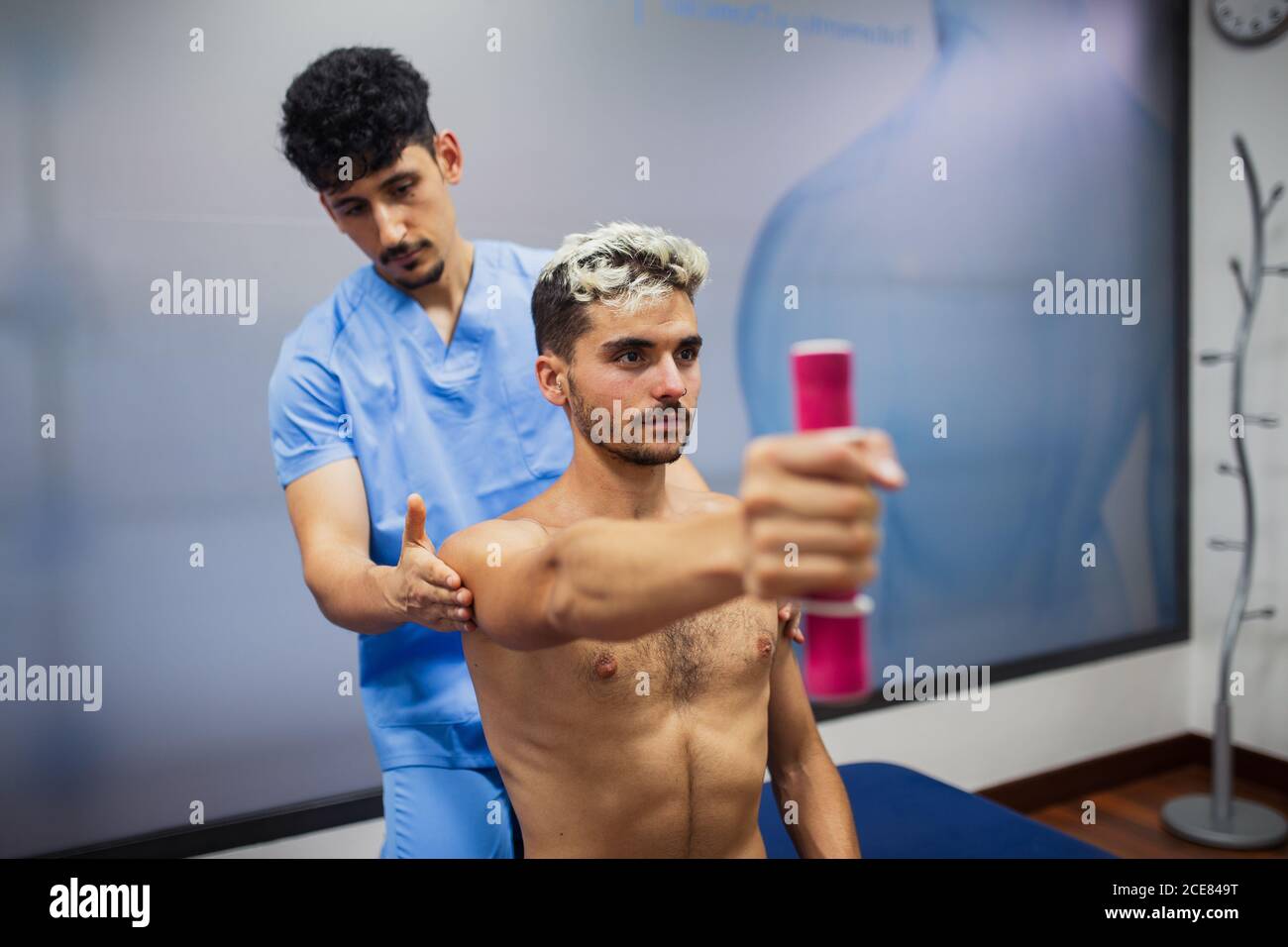 Serious sports medicine osteopath checking up shoulder of calm fit male with dyed hair sitting with reached arm and holding dumbbell during rehabilitation process and looking away Stock Photo