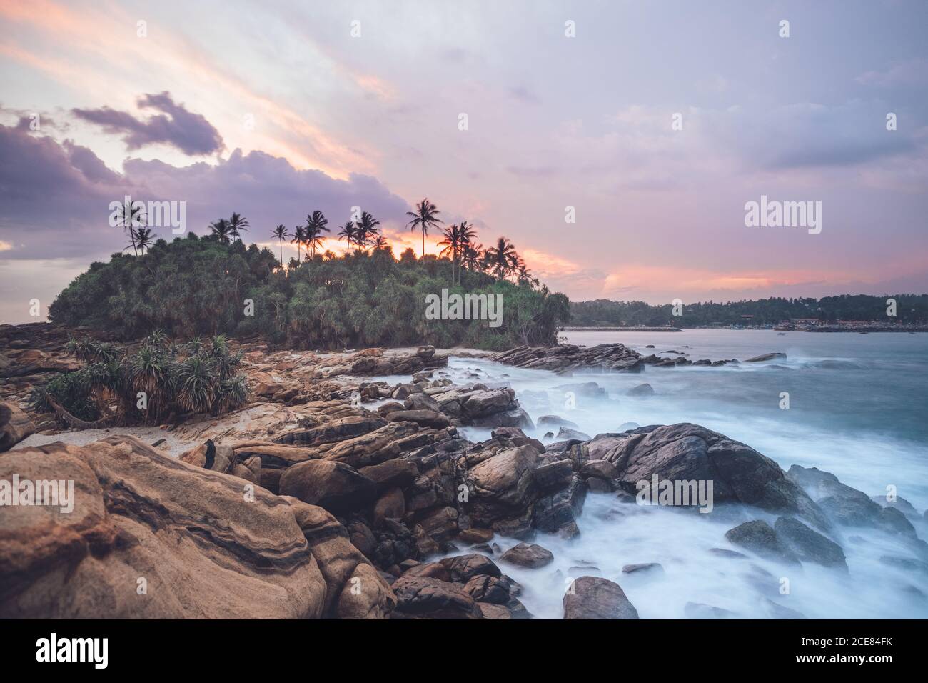 Amazing scenery of rocky coast and green palm trees near sea under picturesque sunset sky Stock Photo