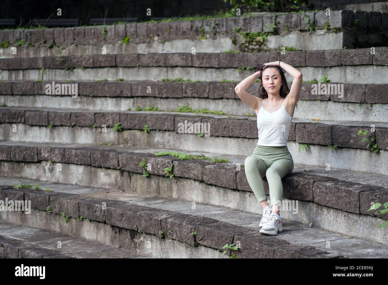 Full length sporty Asian lady wearing tight casual clothes resting on old stone staircase in park Stock Photo