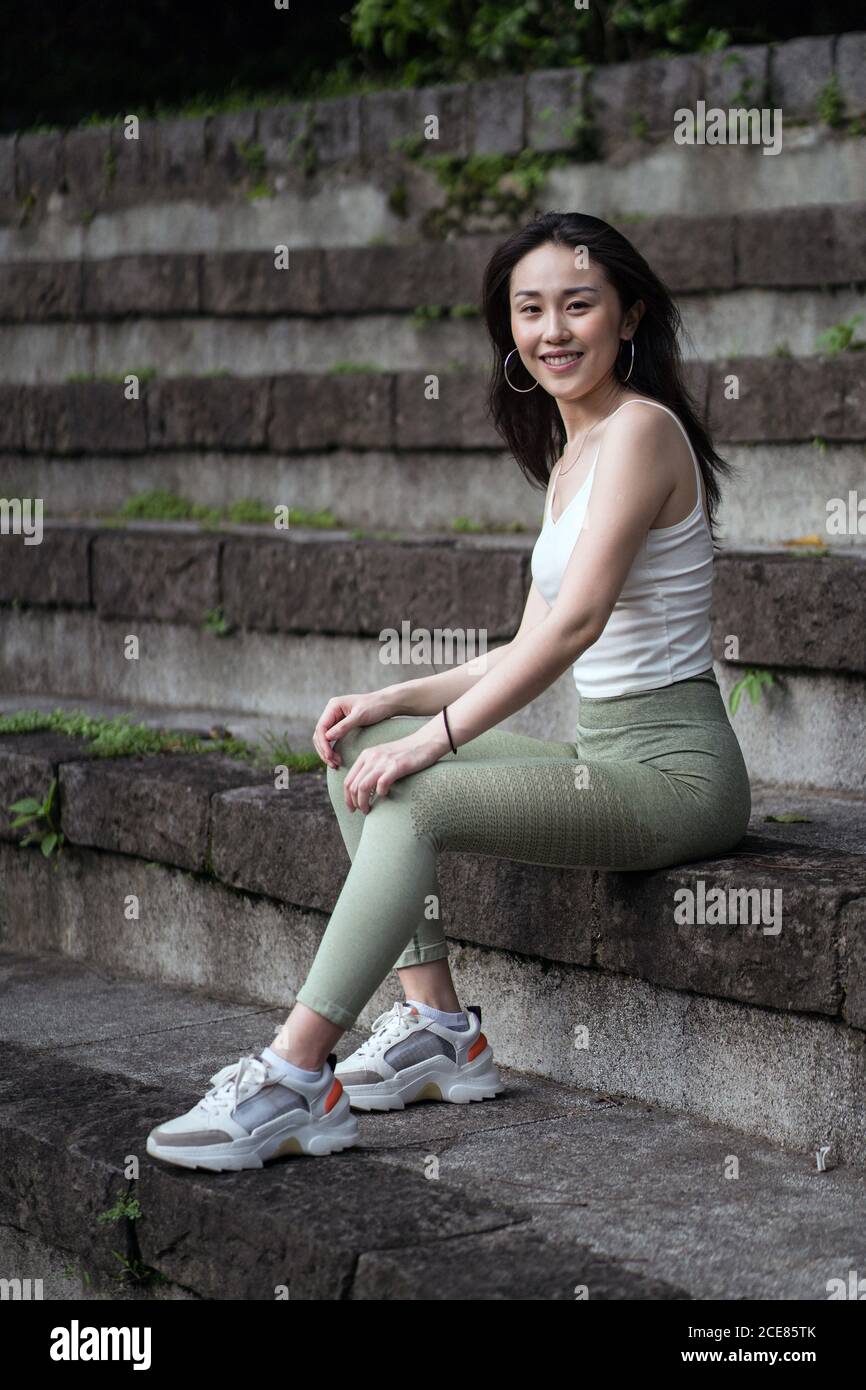 Side view full length sporty Asian lady wearing tight casual clothes resting on old stone staircase in park while looking at camera happily Stock Photo