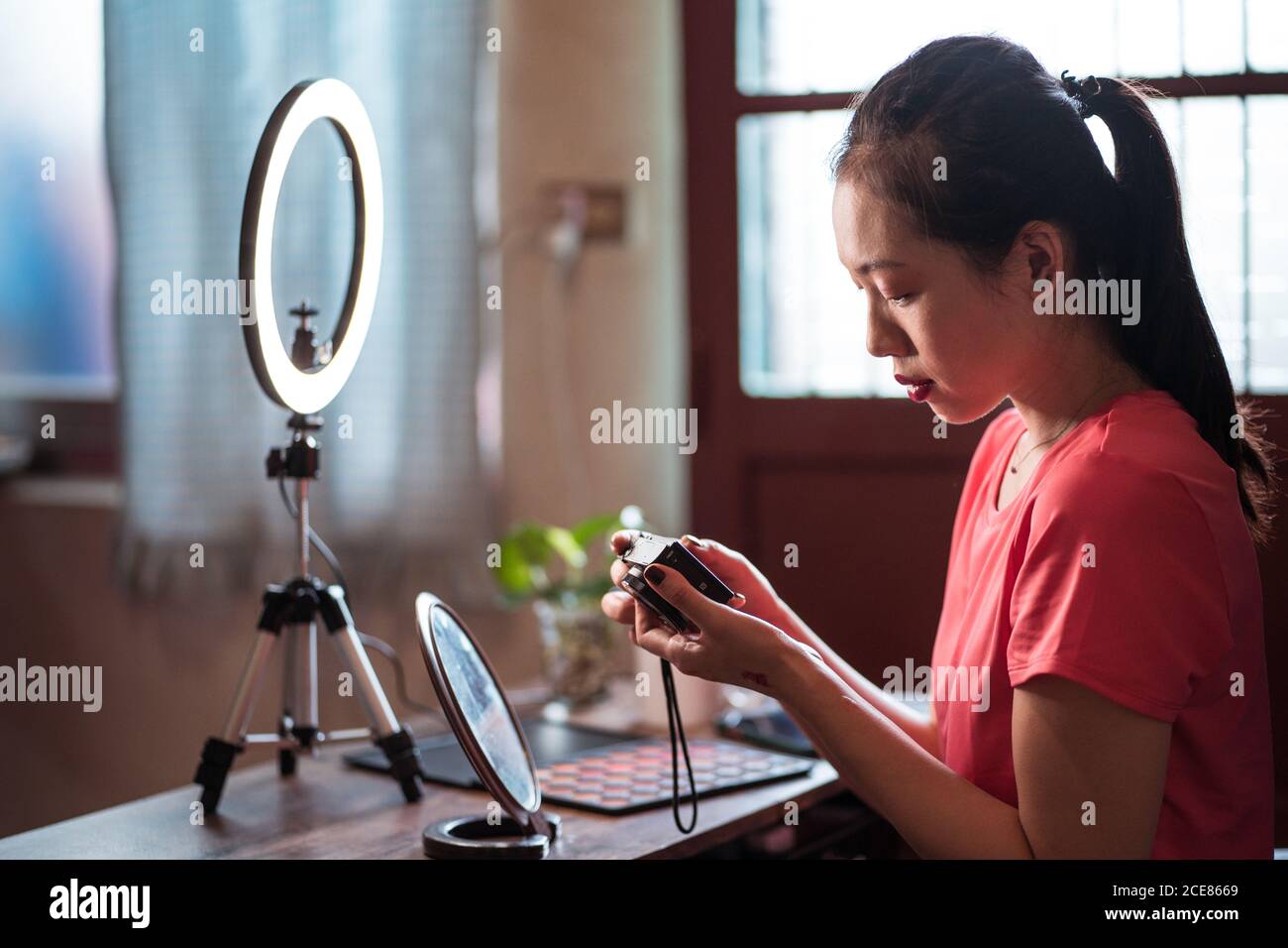 Side view of focused young Asian Woman sitting at table with cosmetics and tools for video shooting and checking video on camera while creating content for beauty blog at home Stock Photo
