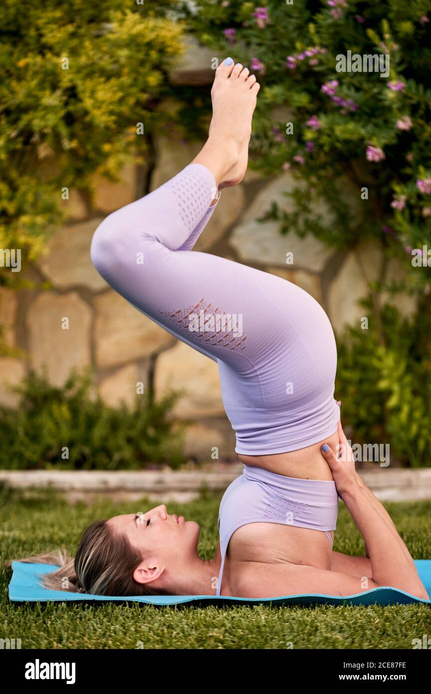 Slim female in bra and leggings practicing yoga in Salamba Sarvangasana on green lawn in backyard in summer Stock Photo