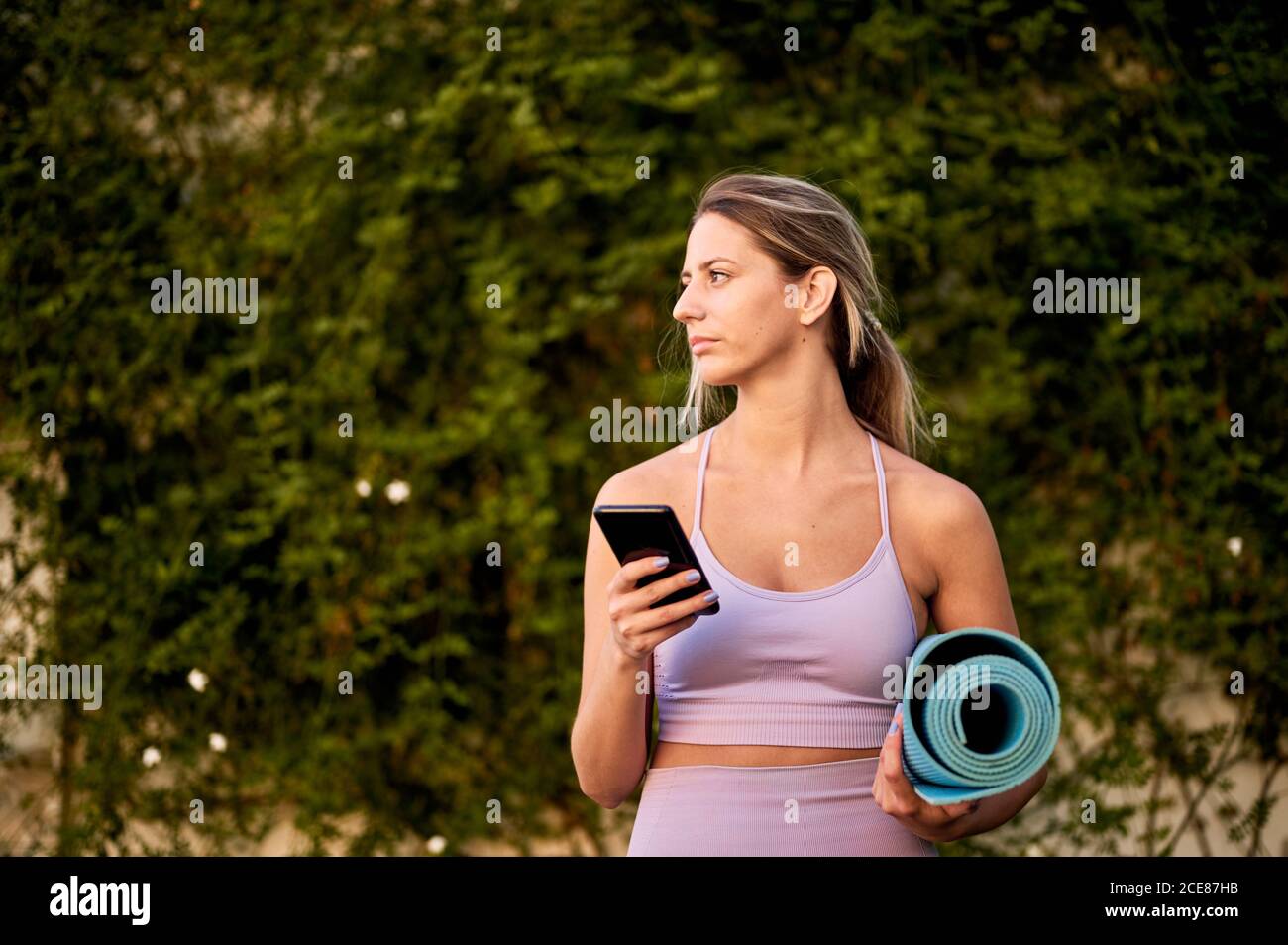 Slim ponder female in sports clothes standing with rolled yoga mat while using cellphone near trees and looking away in daylight Stock Photo