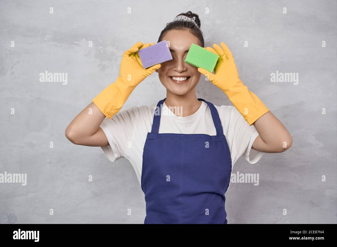 Funny cleaning lady in uniform and rubber gloves covering eyes with kitchen sponges for cleaning and washing dishes, having fun while standing against grey wall. Cleaning services, Housekeeping Stock Photo