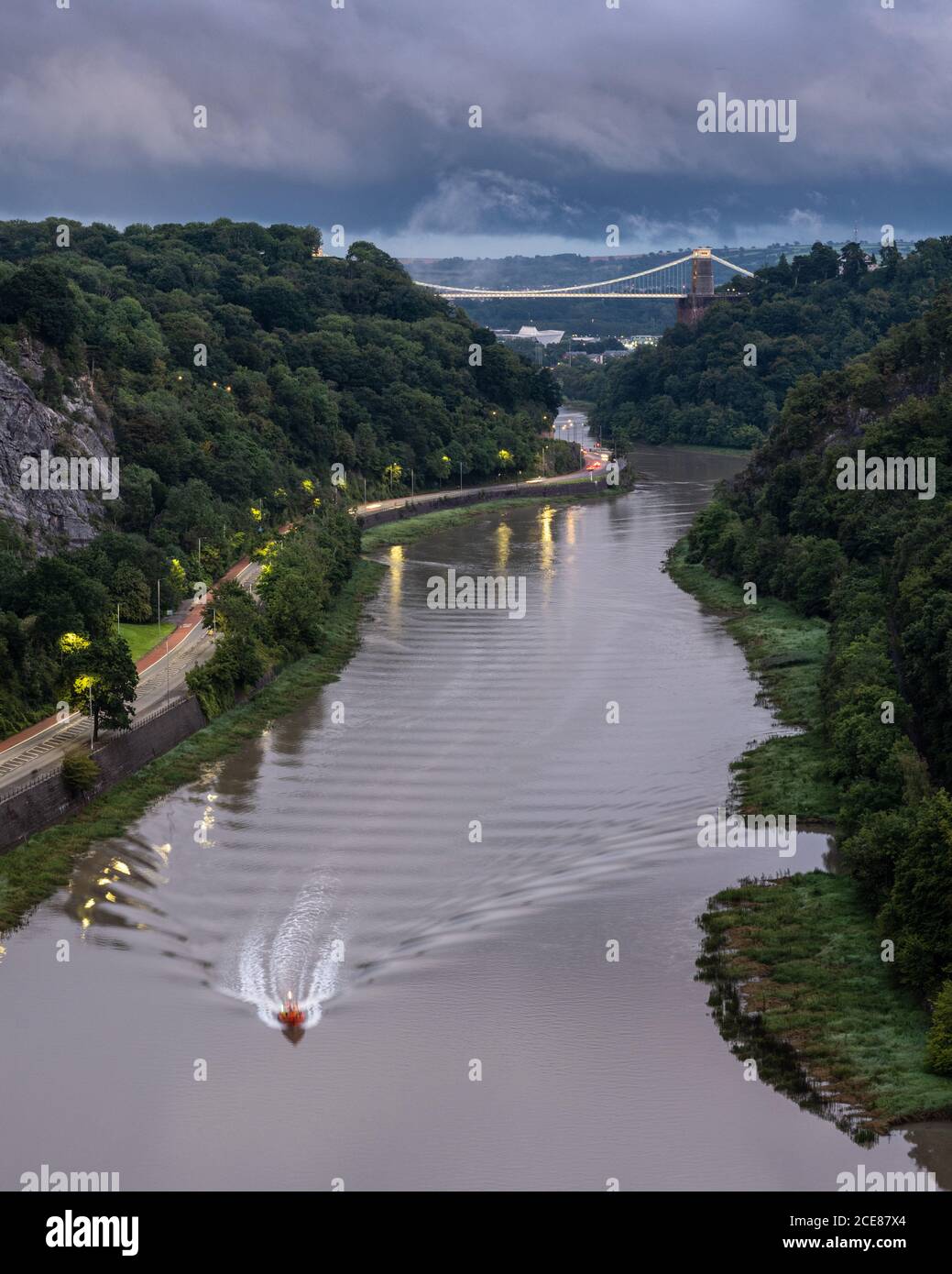 A boat travels down the River Avon estuary at high tide, under the iconic Clifton Suspension Bridge in Bristol's Avon Gorge. Stock Photo