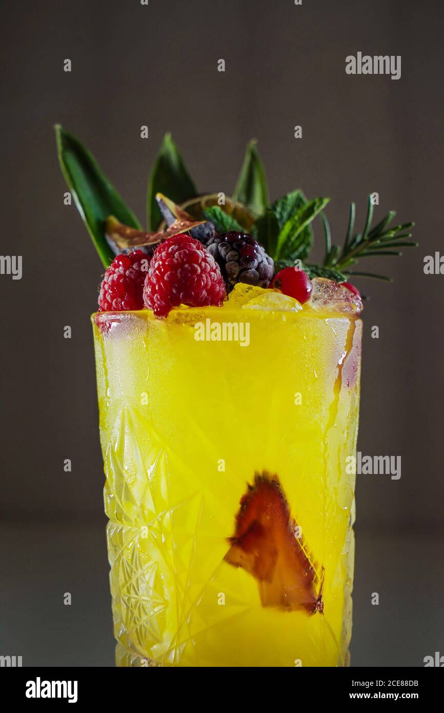 Crystal glass of bright yellow cocktail drink topped with greenery and ripe raspberries on gray background Stock Photo