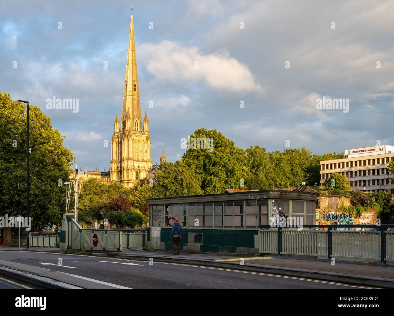 A pedestrian crosses Bristol's Floating Harbour on Redcliffe Bridge, with evening sun lighting the landmark spire of St Mary Redcliffe church behind. Stock Photo