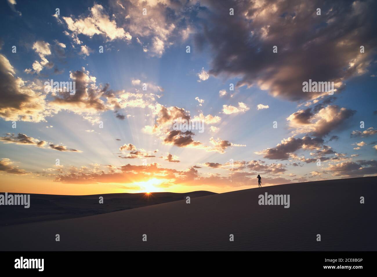 anonymous female tourist strolling in sandy terrain in desert with dry wavy surface under cloudy sky at bright sundown Stock Photo
