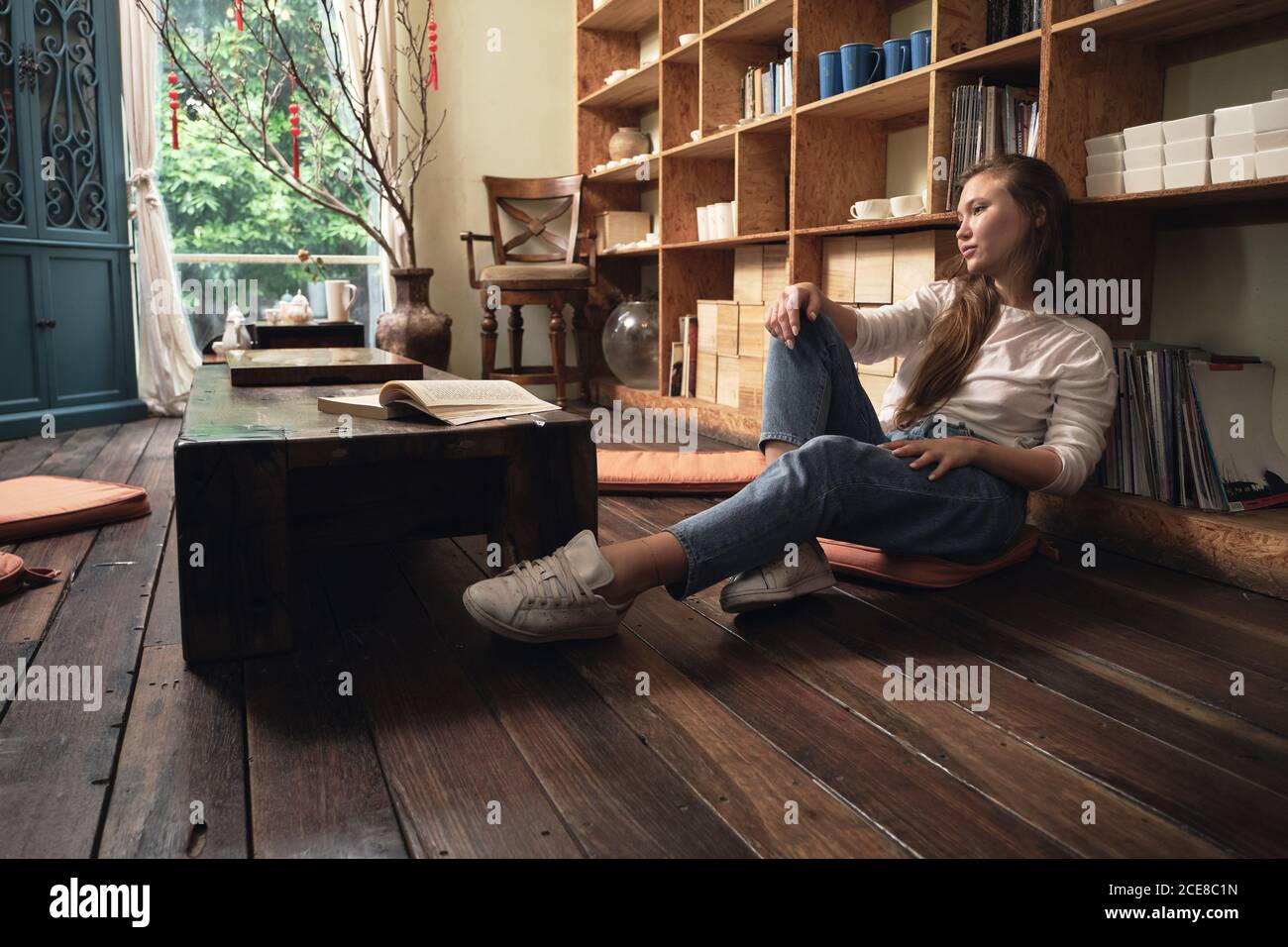 Side view of thoughtful young female student in casual clothes reading textbook while sitting on floor at low table in oriental cafe leaning on a bookcase Stock Photo
