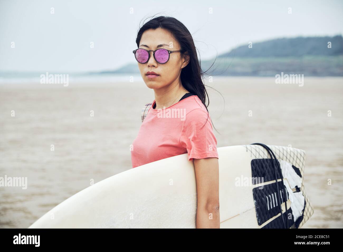 Young Asian female surfer in summer outfit walking on sandy beach and carrying surfboard against calm blue sea Stock Photo
