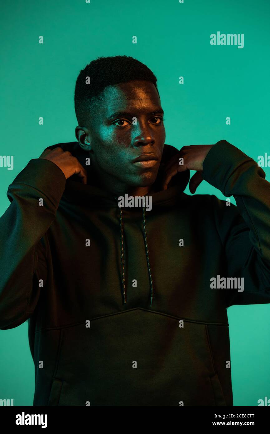 Serious young African American male wearing a black hoodie covering head with hood against green background in studio Stock Photo