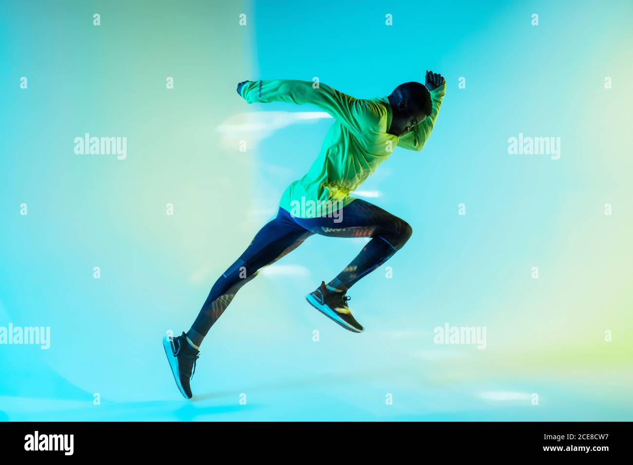 Full body side view of young African American male runner in colorful tracksuit sprinting from start position in studio with bright neon illumination Stock Photo