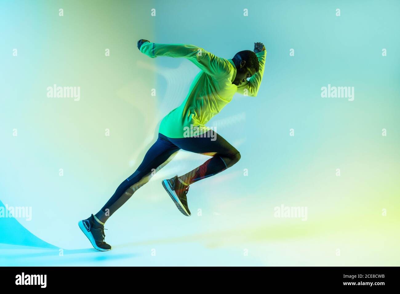 Full body side view of young African American male runner in colorful tracksuit sprinting from start position in studio with bright neon illumination Stock Photo