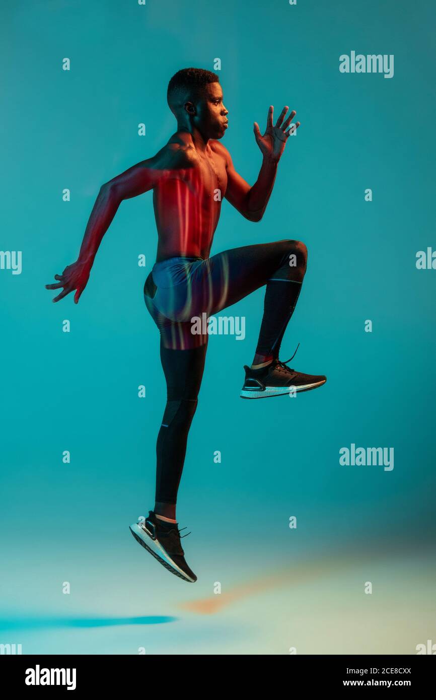 Full body side view of young African American male runner in colorful tracksuit sprinting jumping in studio with bright neon illumination Stock Photo