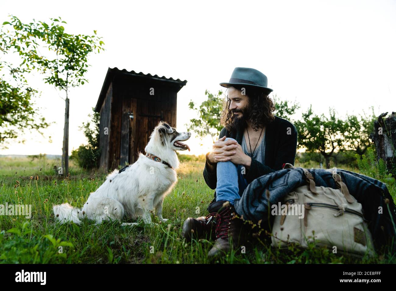 Content male hipster relaxing on grass in field with fluffy dog during sunset in summer Stock Photo