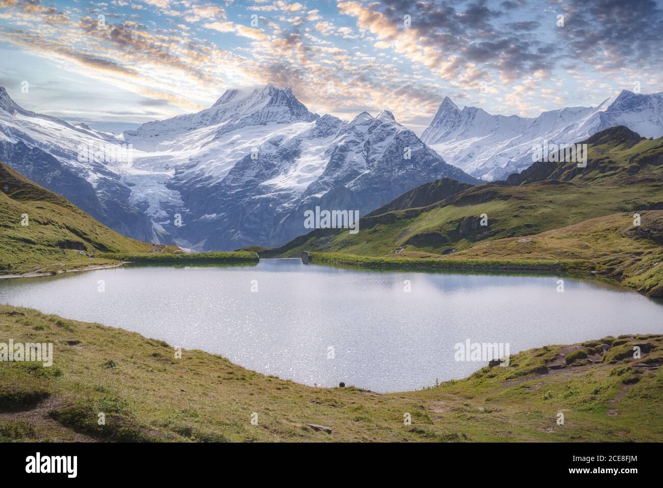 Breathtaking view of small pond with rippled water surrounded by green hills and snowy ridge under blue sky with clouds in afternoon Stock Photo