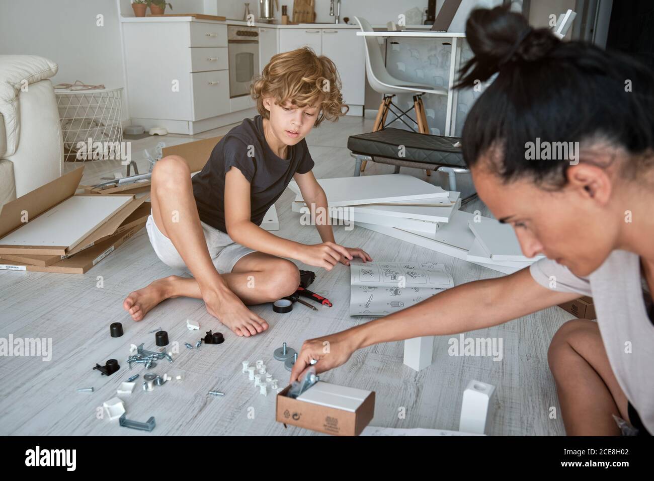 Focused preteen boy sitting on floor with new furniture parts and examining paper instruction while helping cropped mother to fix wardrobe in room with dog lying nearby Stock Photo