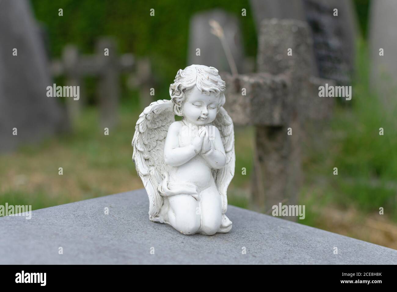 White stone statue of a praying angel in a cemetery with a blurred cross in the background Stock Photo