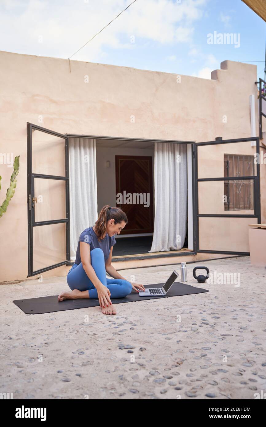 Side view of cheerful female athlete sitting on mat on terrace and picking online tutorial for workout Stock Photo