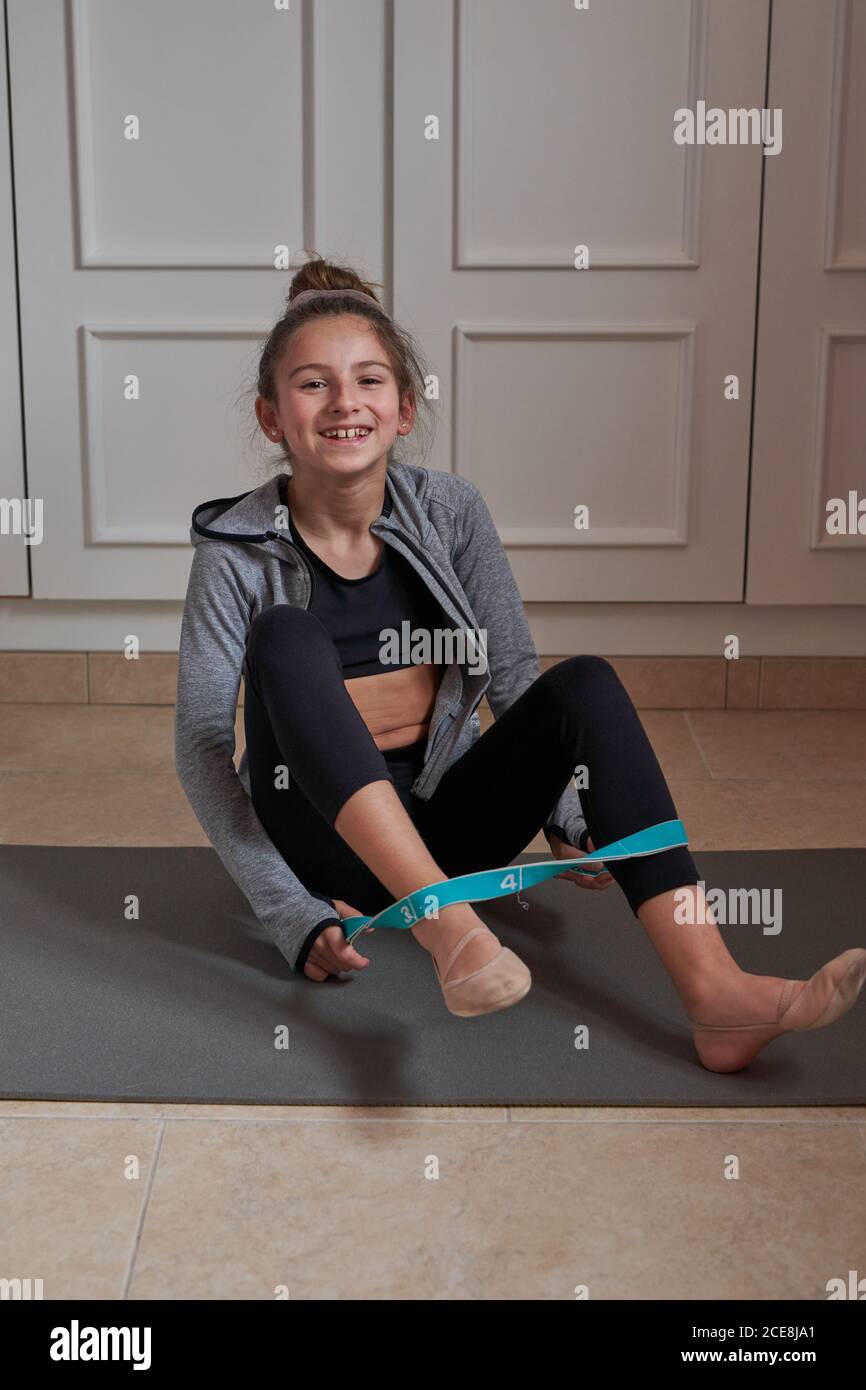 Cheerful teen girl in sportive clothes sitting on mat at home and training  with elastic band smiling at camera Stock Photo - Alamy