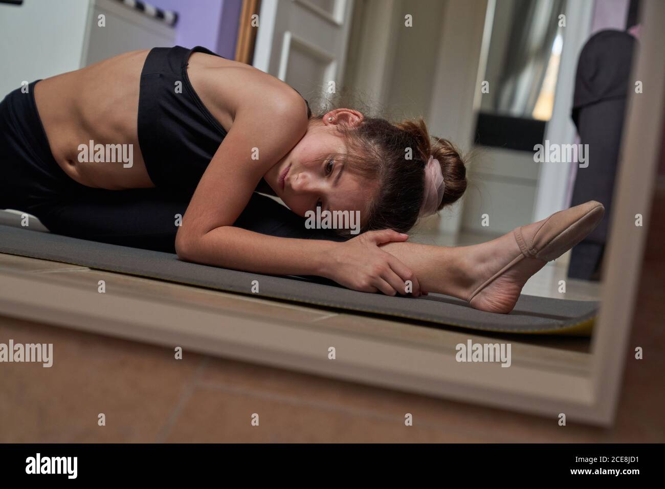 Slim teenage girl sitting on mat doing splits practicing gymnastics in front of mirror Stock Photo