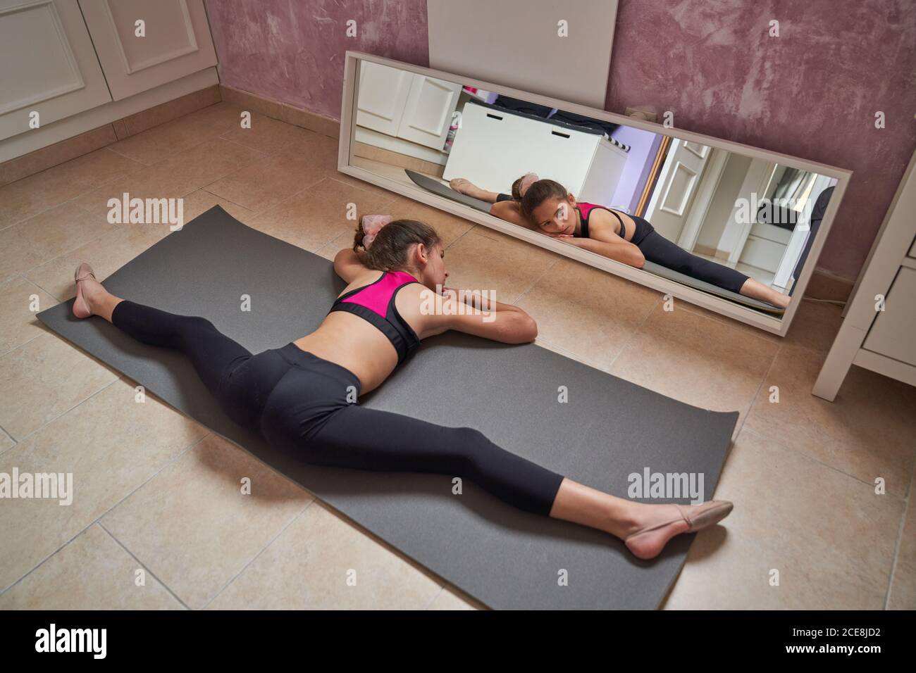From above slim teenage girl sitting on mat doing splits with arms raised practicing gymnastics in front of mirror Stock Photo