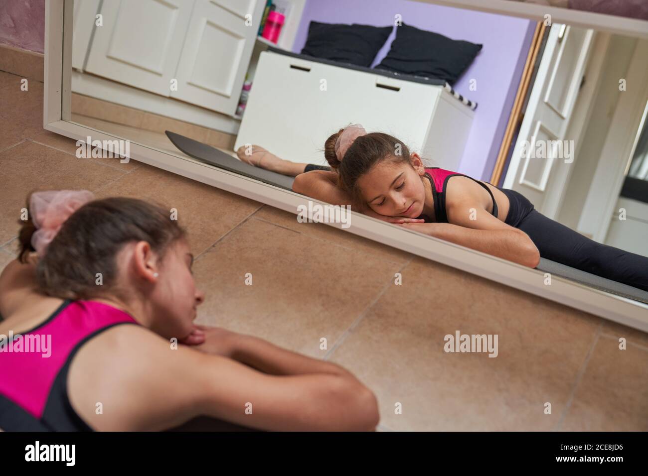 Slim teenage girl sitting on mat doing splits with closed eyes practicing gymnastics in front of mirror Stock Photo