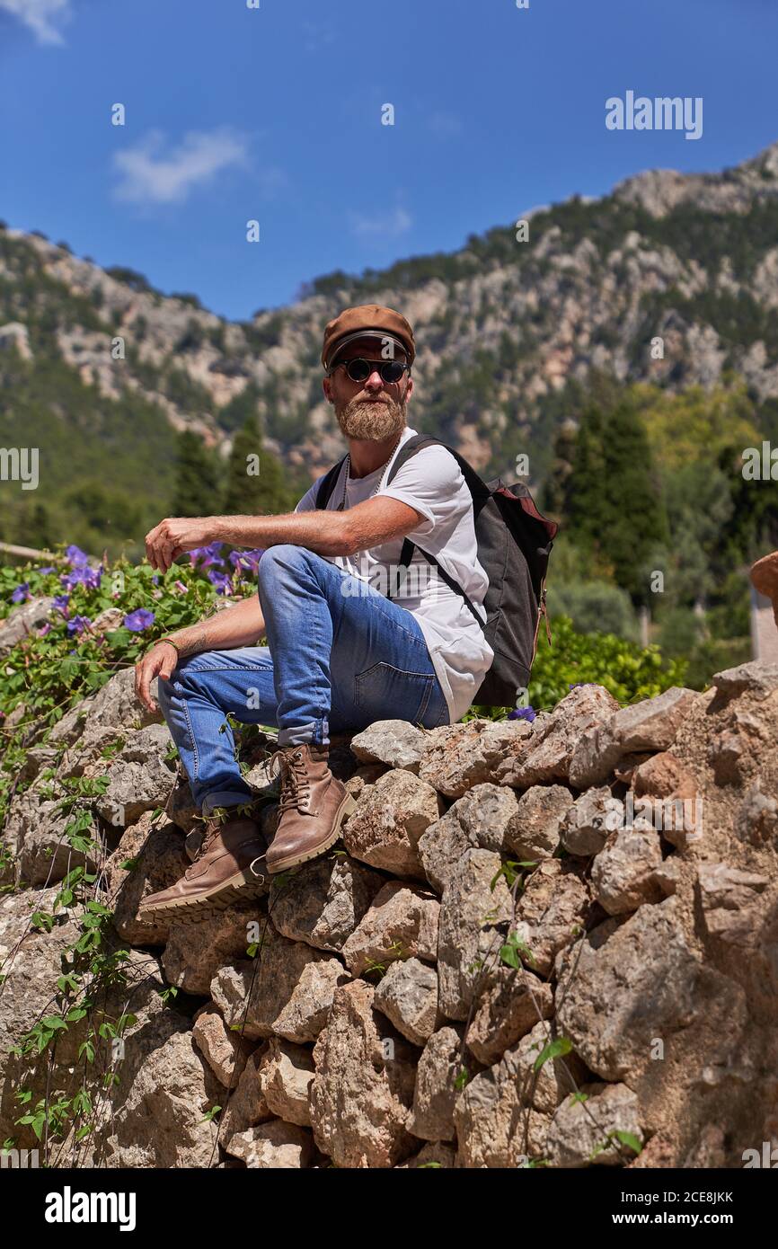 Low angle of calm hipster male backpacker in stylish wear sitting on stony fence against blurred mountains while resting during trekking in summer day Stock Photo