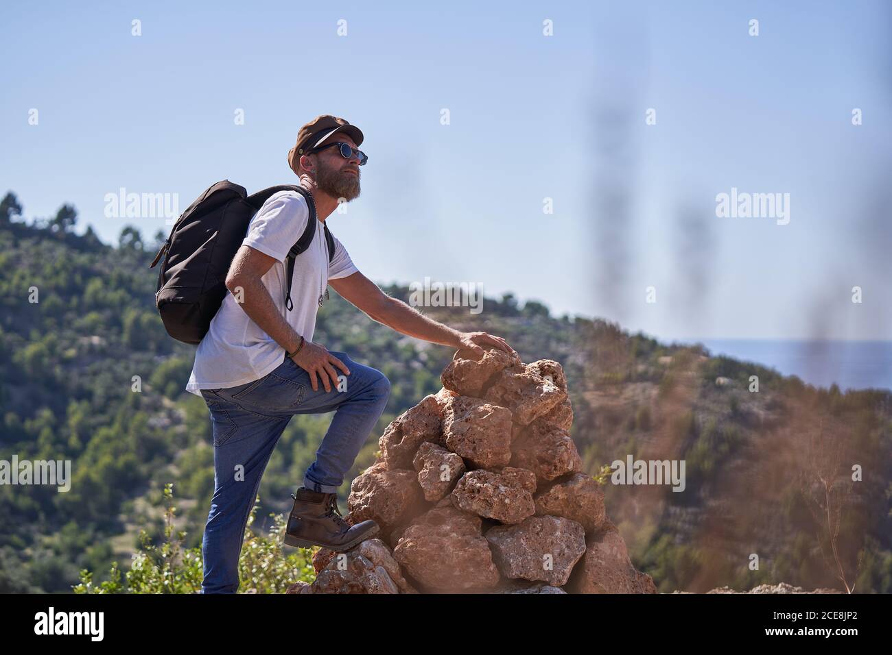 Side view of hipster male backpacker in trendy outfit with sunglasses and cap standing near stone formation and exploring landscape while travelling on coastline in summer Stock Photo