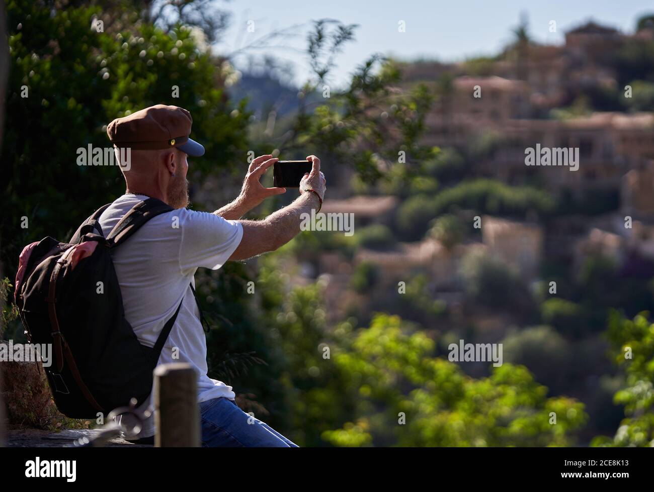 Side view of bearded male backpacker in trendy outfit and cap taking picture on smartphone of ancient stone town located among green hills in summer day Stock Photo