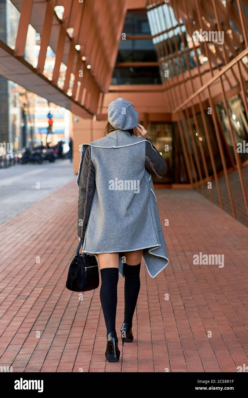 Back view of well dressed female entrepreneur walking along futuristic building and discussing business issues on smartphone Stock Photo