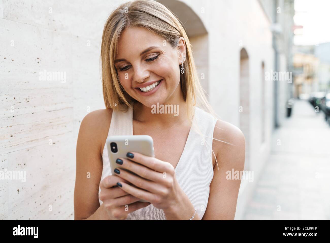 Image of happy blonde woman smiling and using mobile phone while walking on city street Stock Photo