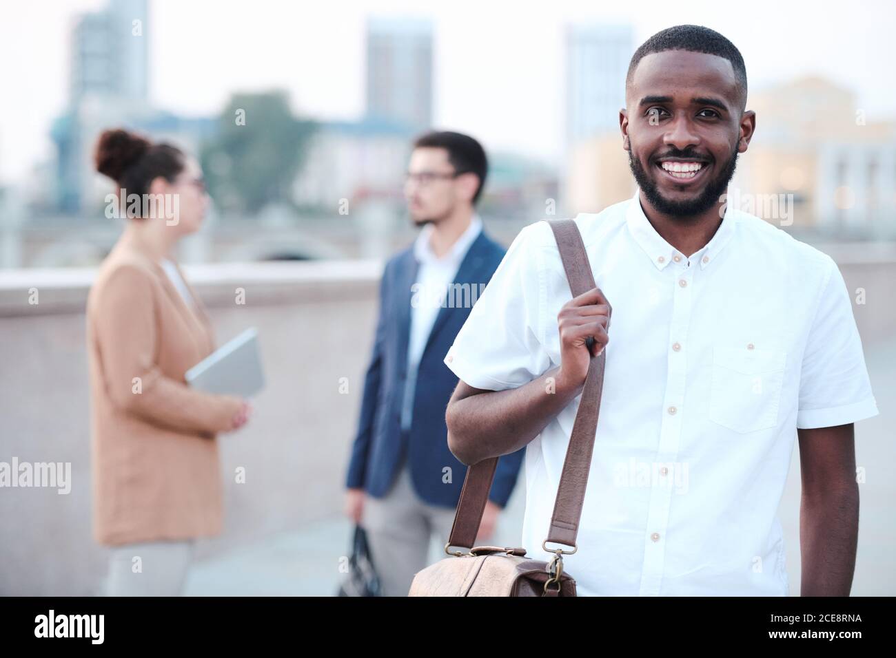 Portrait of cheerful Afro-American student guy with beard holding briefcase outdoors, their group mates talking in background Stock Photo