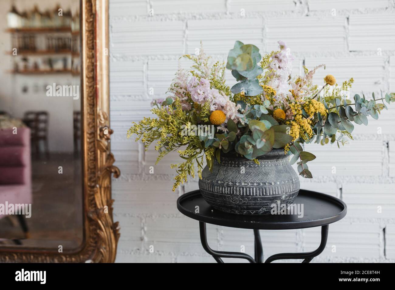 Beautiful bouquet with various flowers including goldenrod and craspedia flowers with green eucalyptus branches arranged in ornamental ceramic pot placed on small table against white brick wall with mirror in creative floristry studio Stock Photo