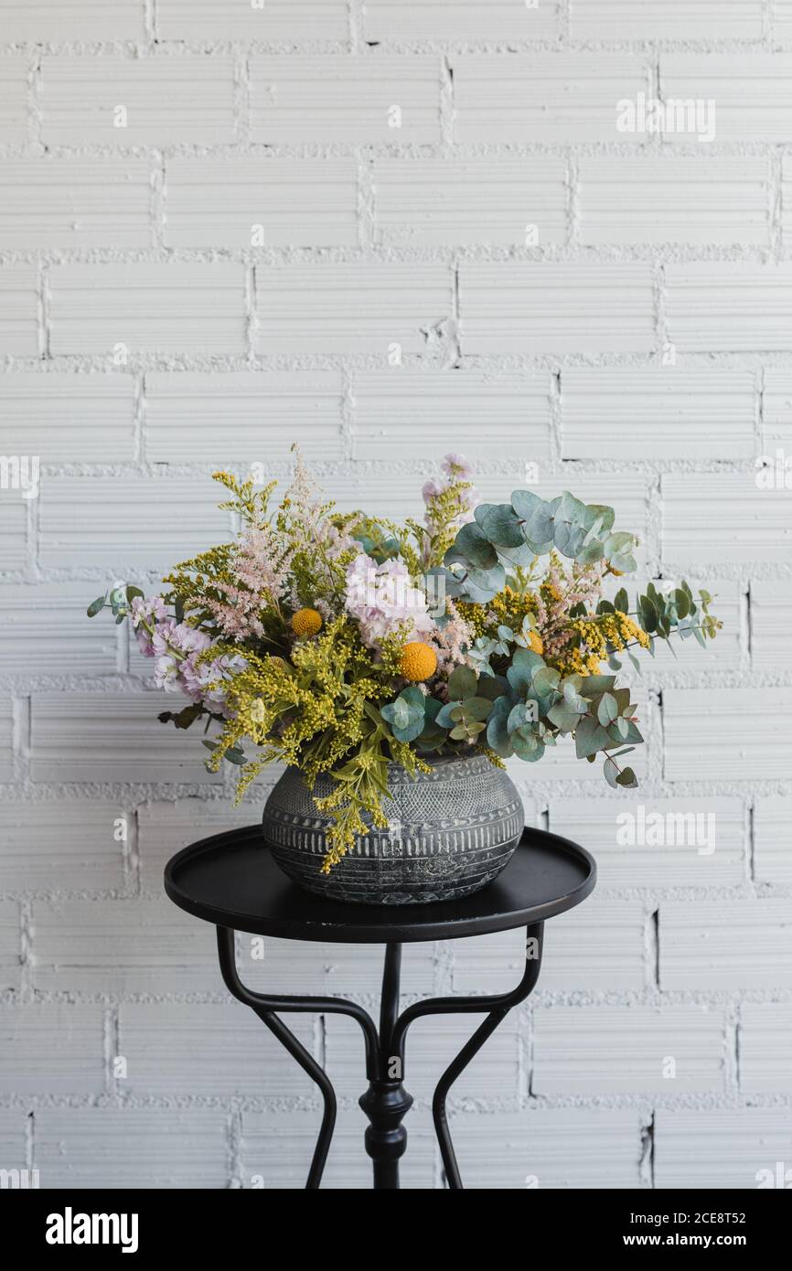 Beautiful bouquet with various flowers including goldenrod and craspedia flowers with green eucalyptus branches arranged in ornamental ceramic pot placed on small table against white brick wall in creative floristry studio Stock Photo