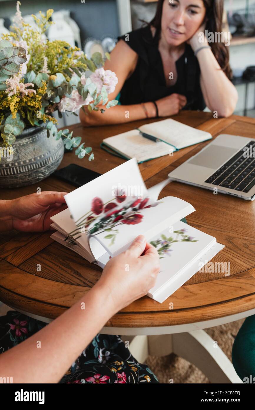Crop anonymous female florist leafing through catalog of decorative plants while sitting at table with colleague and discussing new order for floristry workshop Stock Photo