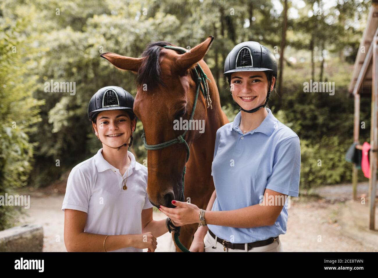 Young smiling female jockeys in helmets standing near horse in countryside and looking at camera Stock Photo