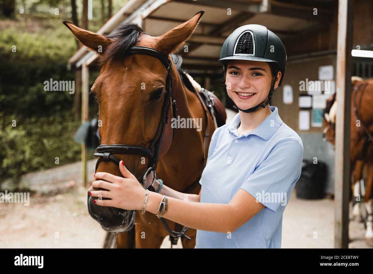 Young smiling female jockey in helmet standing near horse in countryside and looking at camera Stock Photo