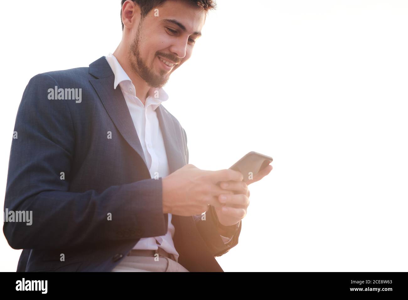 Below view of smiling bearded manager in jacket using mobile internet on phone outdoors Stock Photo