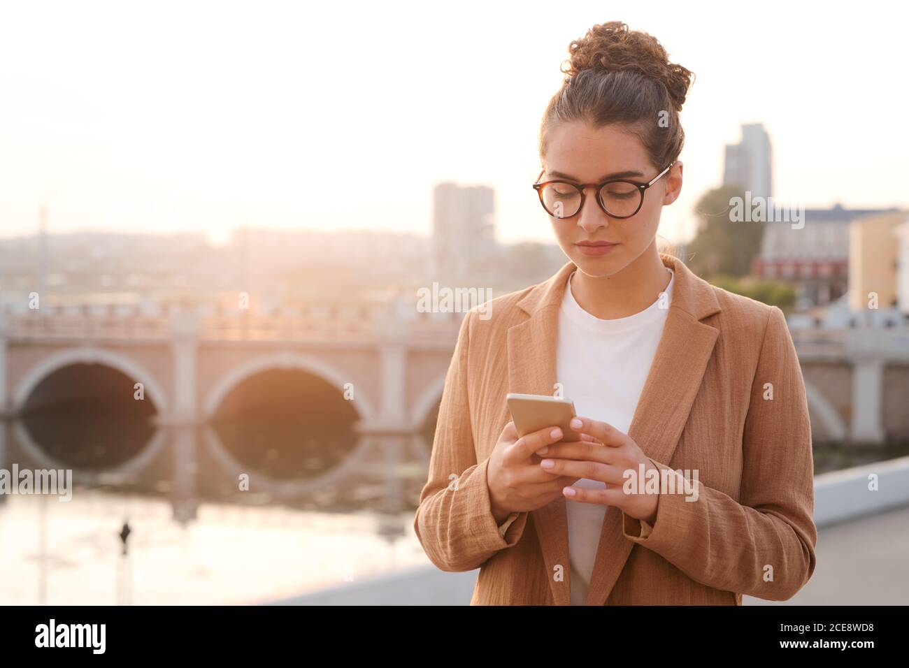 Serious young businesswoman with hair bun checking messengers on smartphone while walking over street Stock Photo