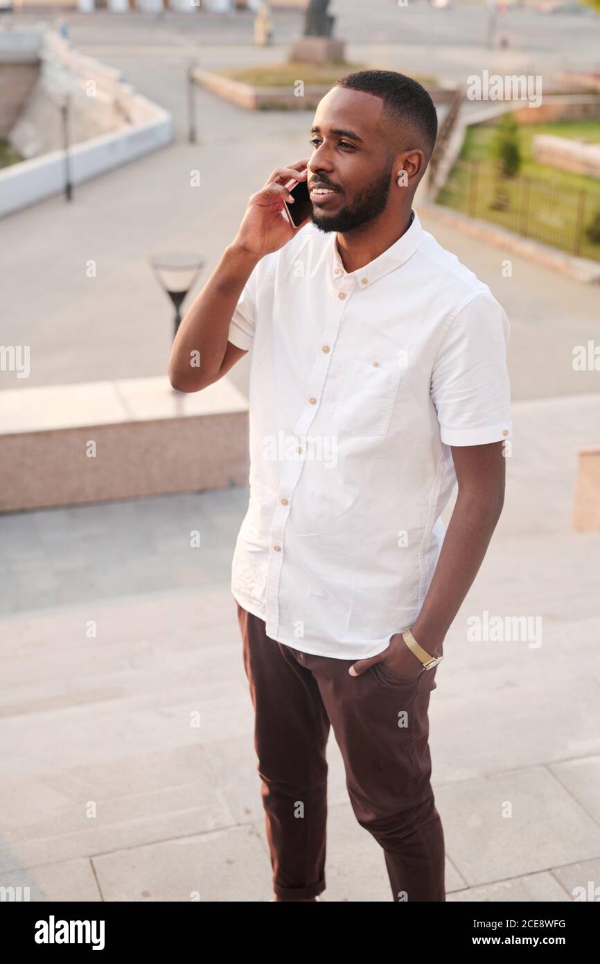 Content young Black manager in white shirt standing outdoors and holding hand in pocket while answering phone Stock Photo