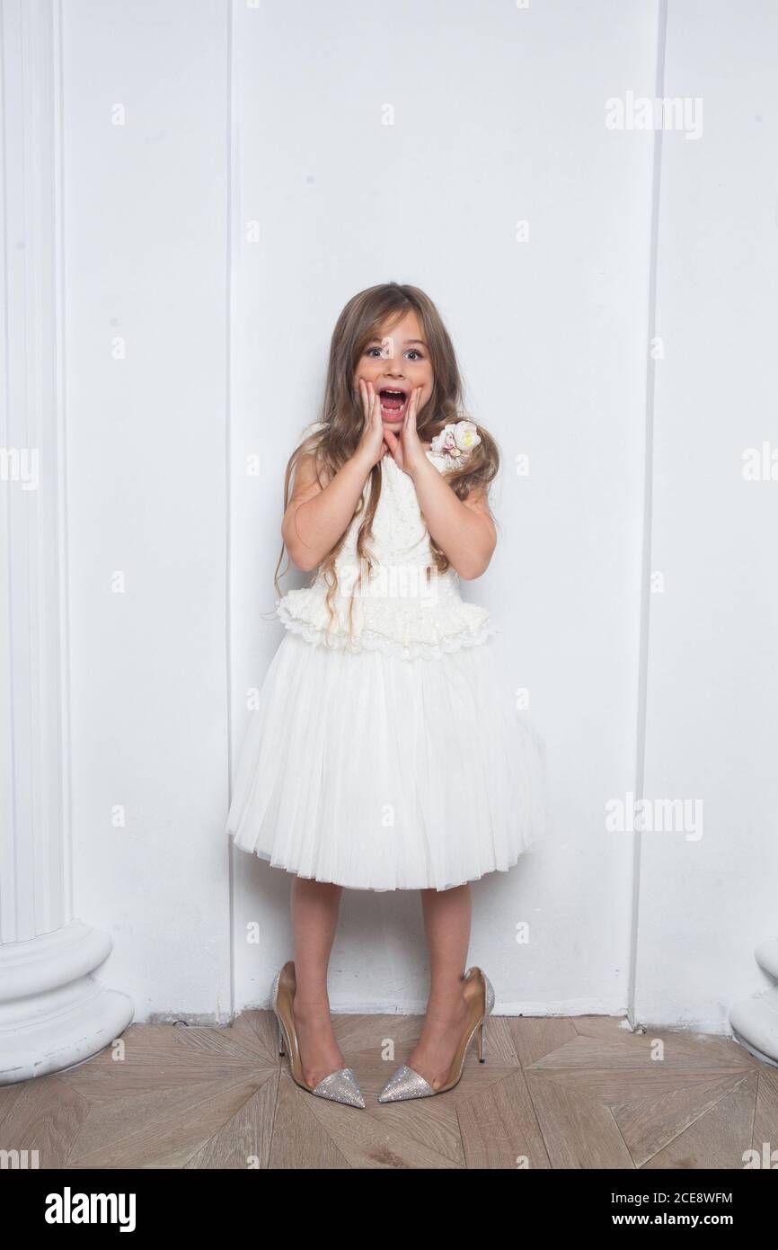 Little princess - excited emotional cute girl in fashion white dress having fun and wearing big mothers sparkle high heels shoes on white background Stock Photo