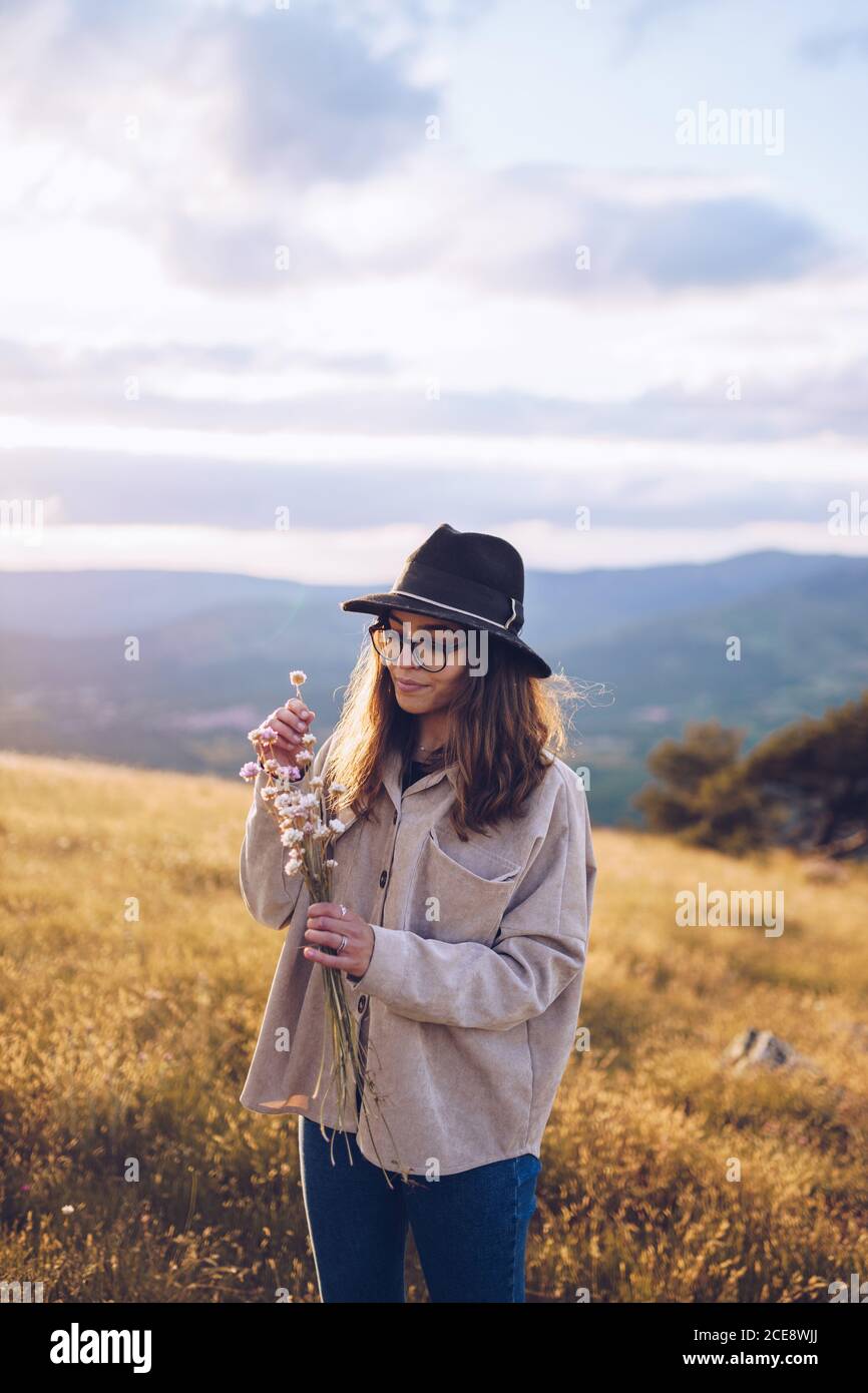 Tender female with bouquet of wildflowers standing on hill on background of majestic mountainous landscape in autumn during sundown Stock Photo