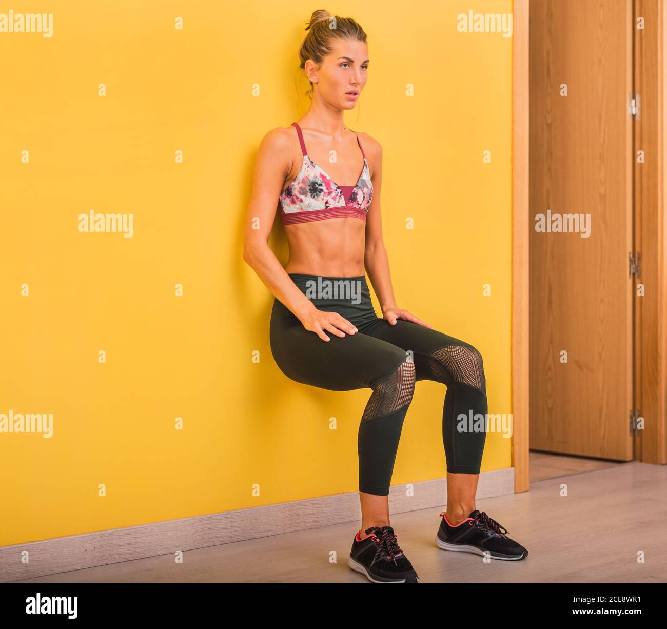 Slim young ponder female in sports clothes sitting with open mouth in Chair pose leaned on bright yellow wall during warm up Stock Photo
