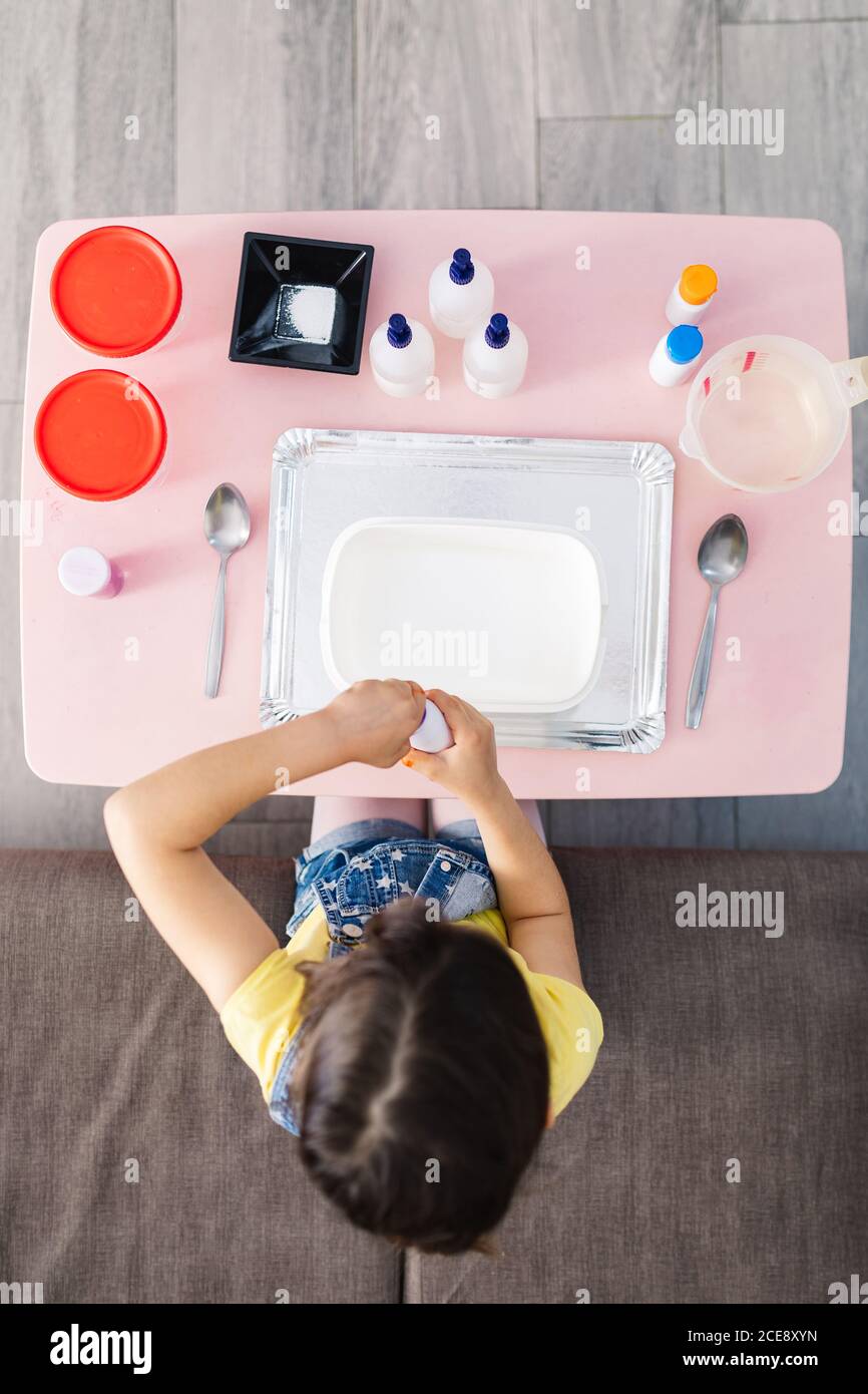 From above of child sitting on comfortable couch while taking plastic bottle with colorant on table with tray and container near spoons Stock Photo