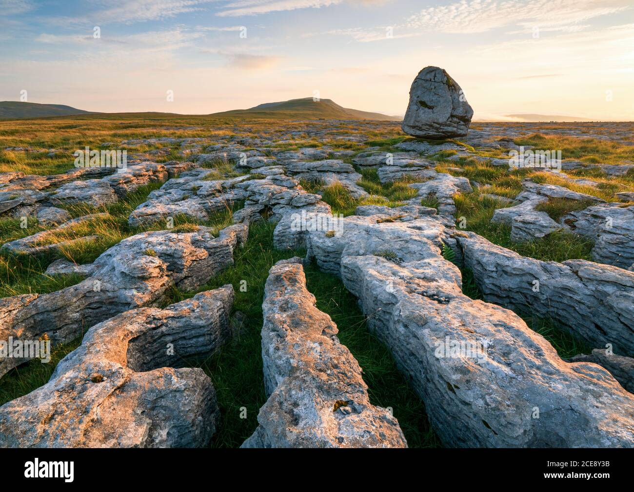 Sinous limestone pavement leads the eye towards Whernside and a distinctive erratic boulder on Scales Moor at sunrise on a blustery summer morning. Stock Photo