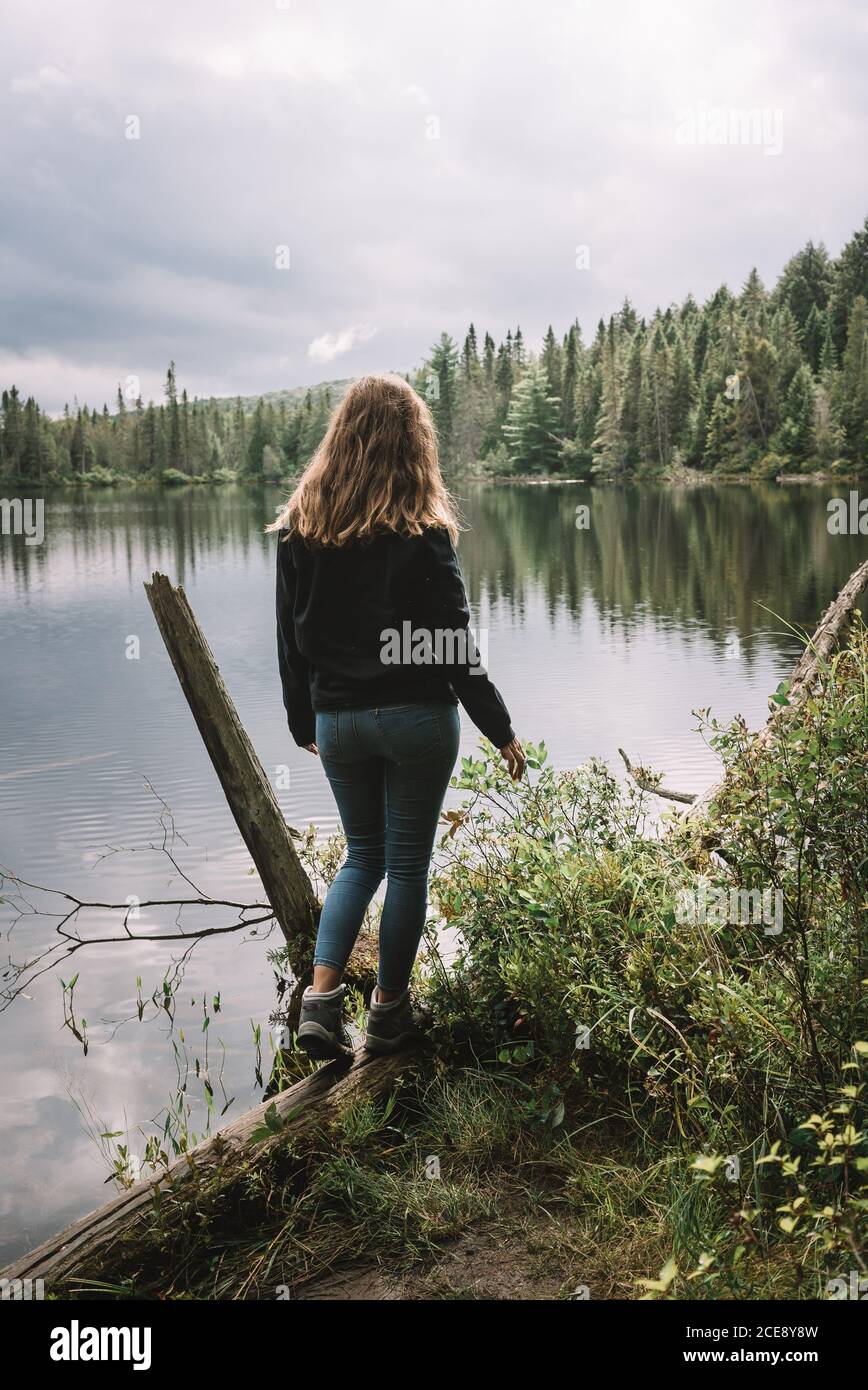 Back view of unrecognizable female tourist in casual outfit standing on  lake shore surrounded by green