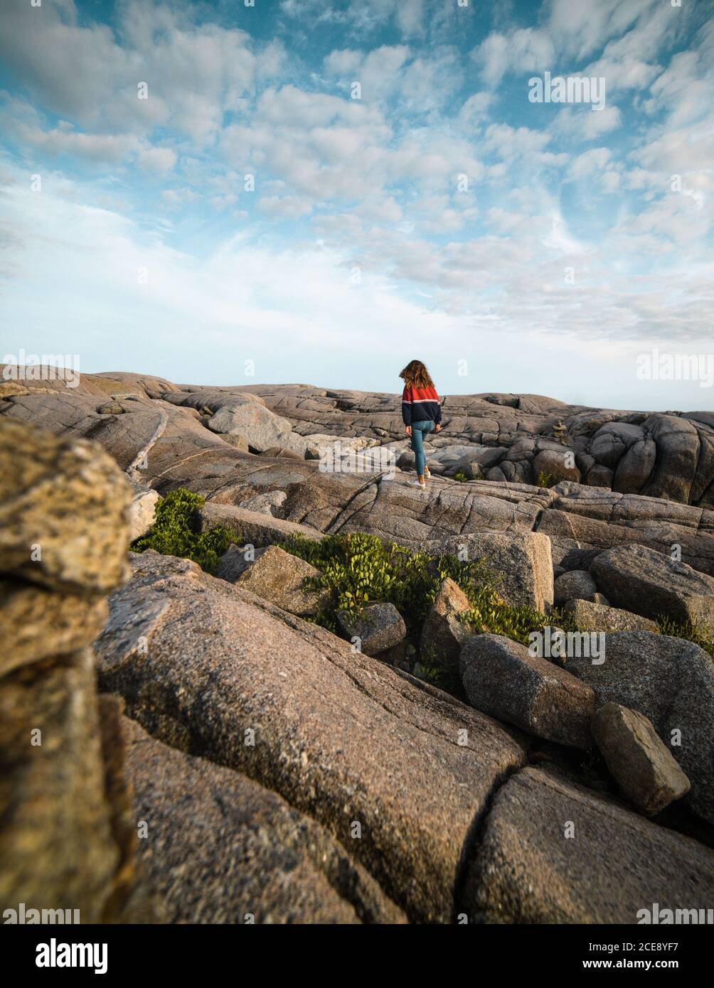 Back view of anonymous female tourist walking on rough stony coast against gray cloudy sky near sea in Peggys Cove, Canada Stock Photo